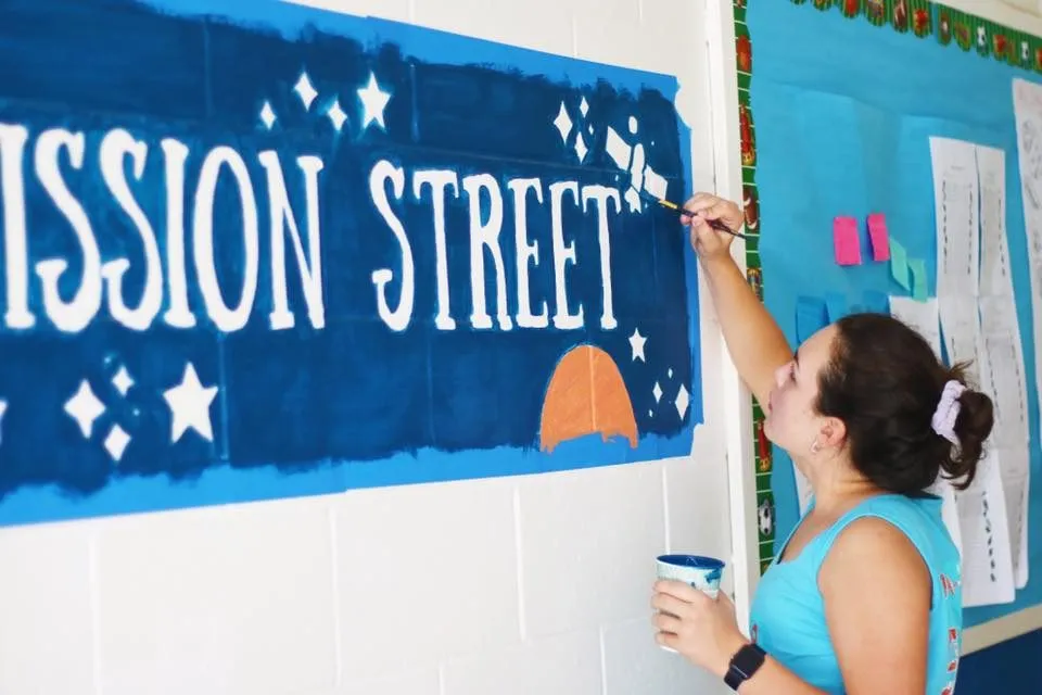 A volunteer painting a night sky mural lettered Mission Street on a corridor wall