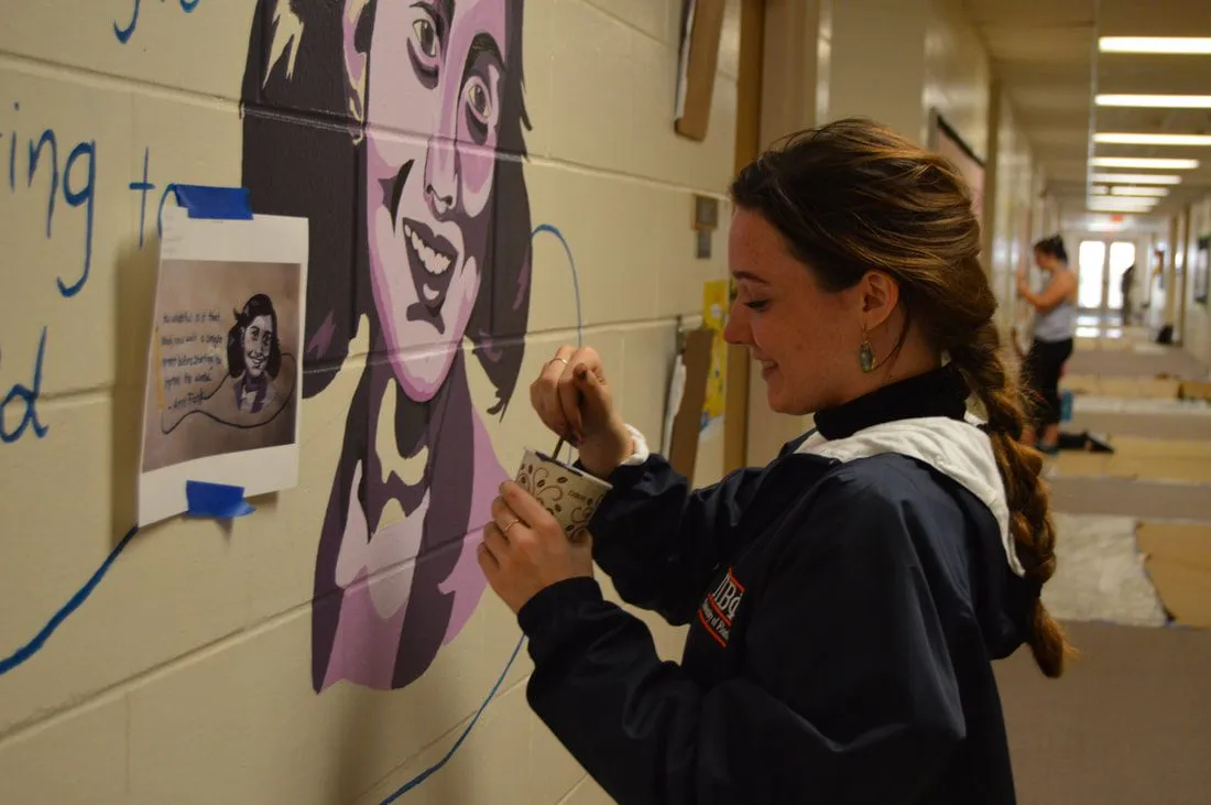 A volunteer painting the Anne Frank portrait mural, working from a taped-up reference photo